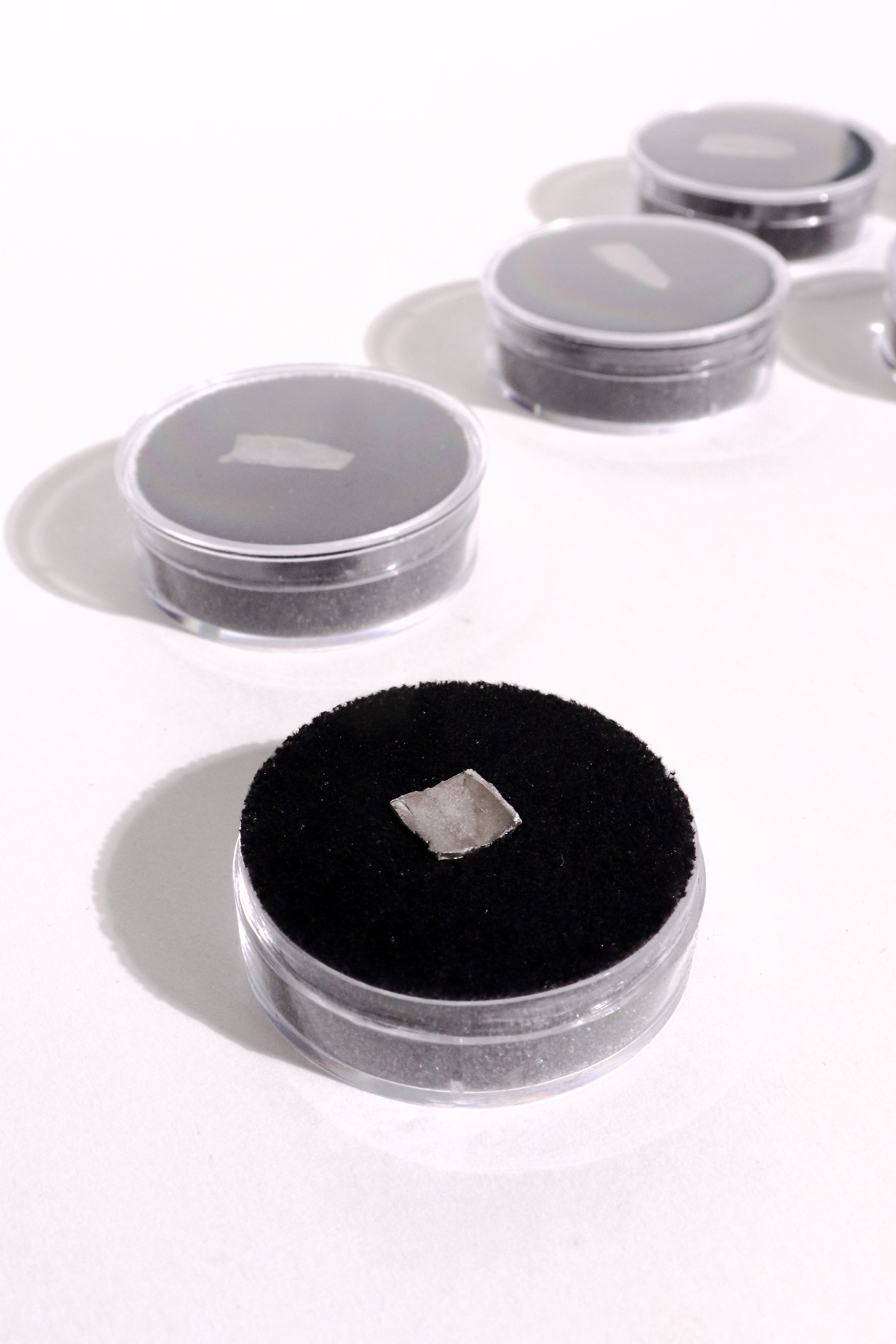 A small square silver-colored piece displayed on a black circular velvet pad inside a clear round container, with several similar containers arranged in the background on a white surface.