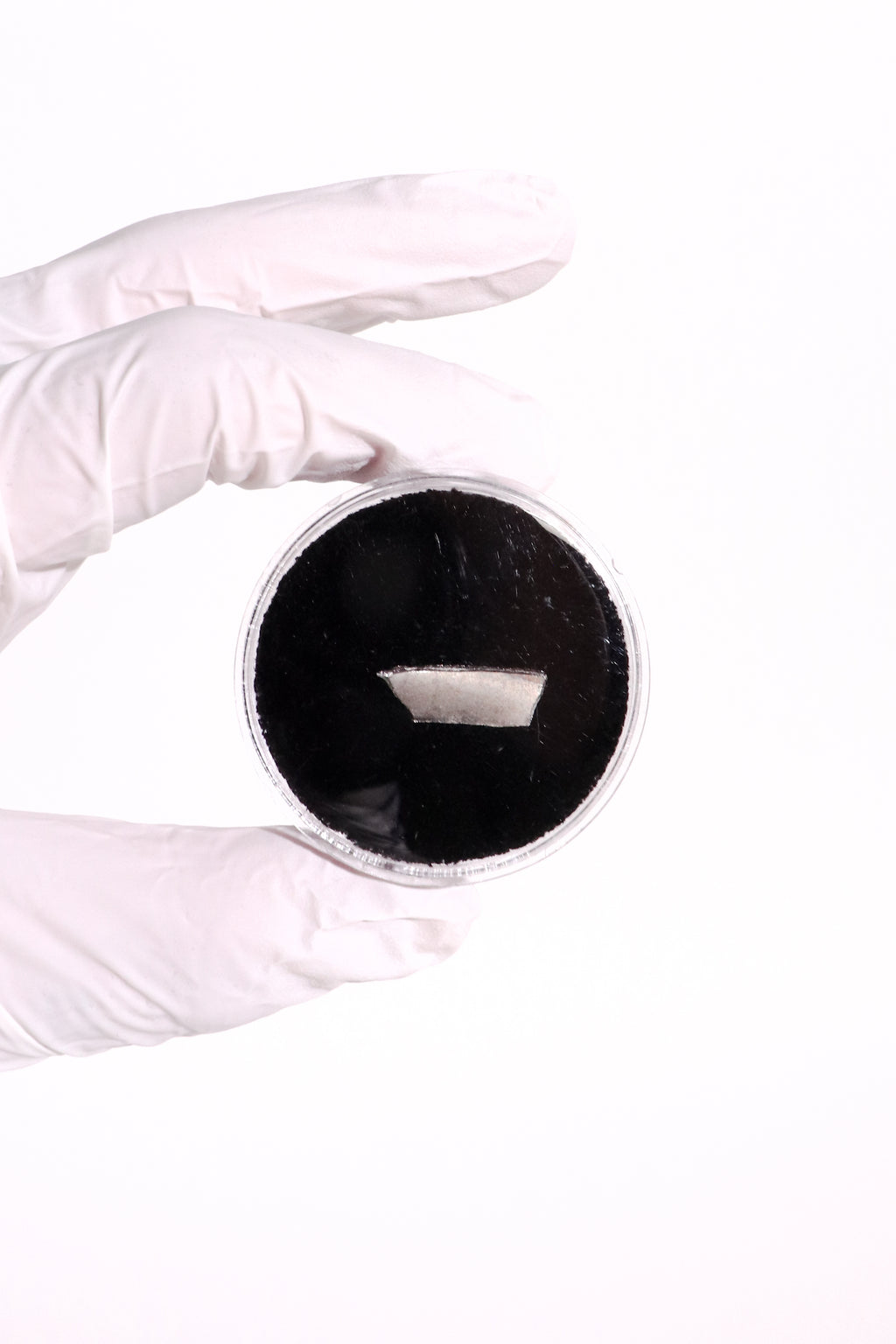 A white-gloved hand holding a small round container filled with a very dark black powder or material against a plain white background.