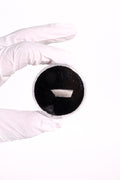 A white-gloved hand holding a small round container filled with a very dark black powder or material against a plain white background.