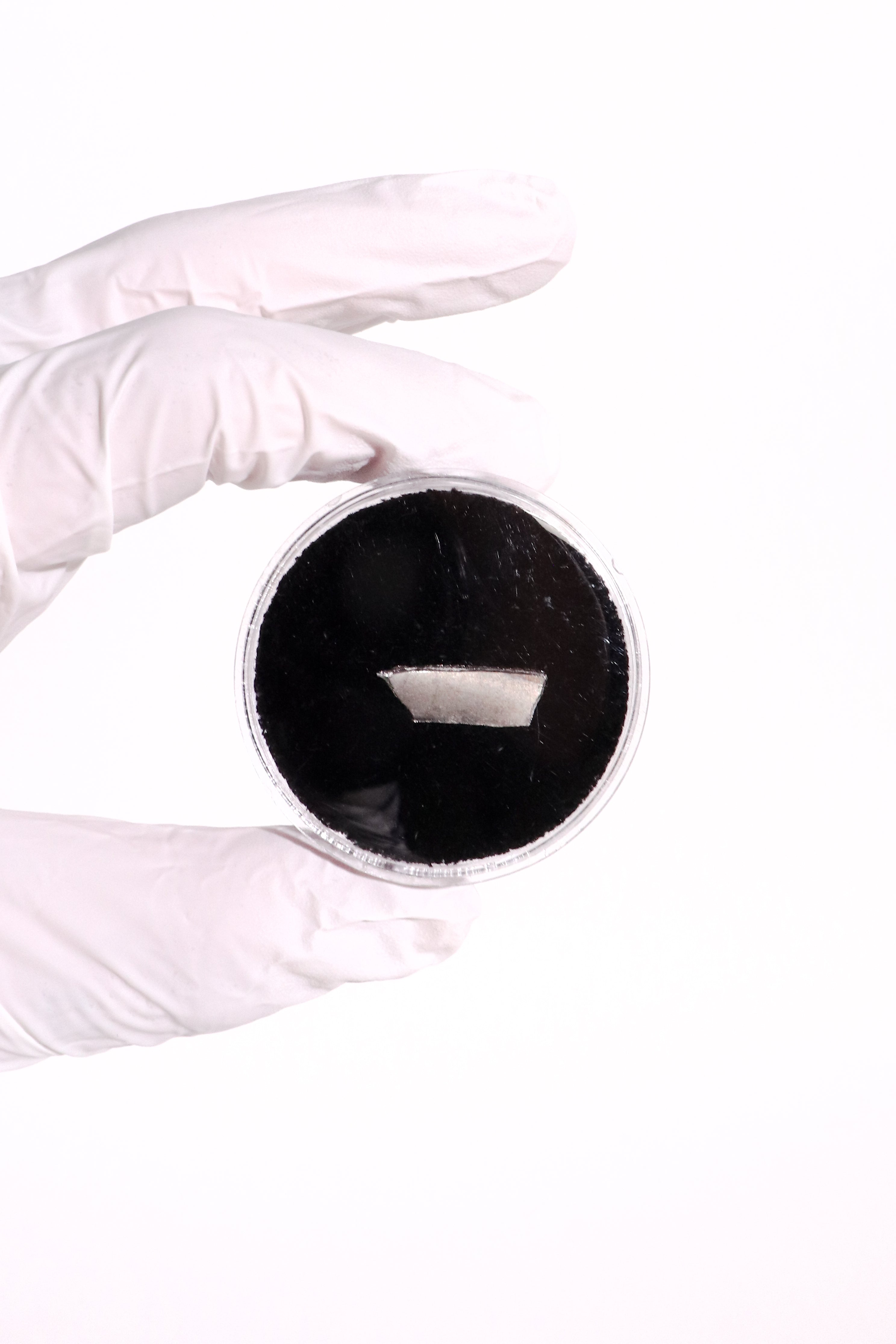 A white-gloved hand holding a small round container filled with a very dark black powder or material against a plain white background.