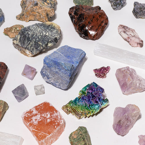 A collection of raw, rough gemstones and crystals in various colors and shapes, arranged on a plain white background. The stones include shades of blue, purple, pink, green, orange, brown, and gray, with some showing a metallic or iridescent sheen.